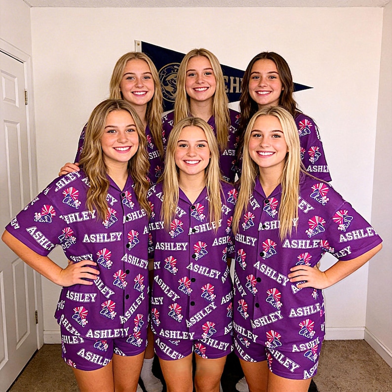 K&ouml;nnte beinhalten: Sechs Personen tragen passende lila Pyjama-Sets. Die Pyjamas haben ein sich wiederholendes Muster mit dem Namen "ASHLEY" und einer Grafik. Die Sets umfassen ein Hemd mit Kn&ouml;pfen und Shorts.