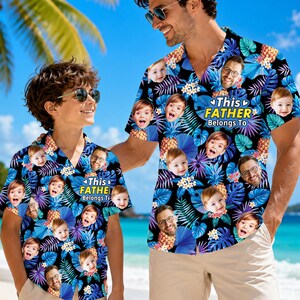 May include: Two short-sleeved button-down shirts featuring a tropical pattern of blue and purple palm leaves, pineapples, and white flowers. The shirts are printed with multiple photos of a child's face and the text This FATHER Belongs To.