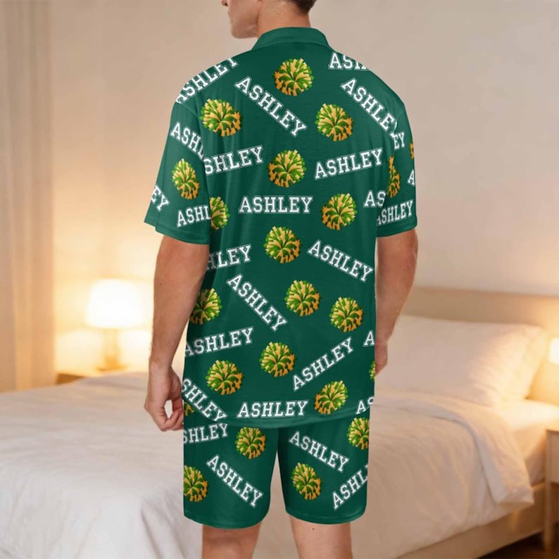 K&ouml;nnte beinhalten: Gr&uuml;nes Pyjama-Set mit einem sich wiederholenden Muster des Namens "ASHLEY" in Wei&szlig; und gelben Pompon-Grafiken. Das Set beinhaltet ein kurz&auml;rmeliges Hemd mit Kragen und passende Shorts.