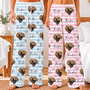 May include: Two pairs of pajama pants featuring a repeating pattern of a golden retriever's face, the name Tucker in script, and paw prints. One pair has light blue and white horizontal stripes, and the other has light pink and white stripes.