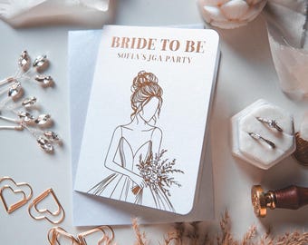 May include: A white card with the text "BRIDE TO BE" and "SOFIA'S JGA PARTY" in gold lettering. The card features a line drawing of a bride holding a bouquet. The card is placed on a white surface with wedding accessories.