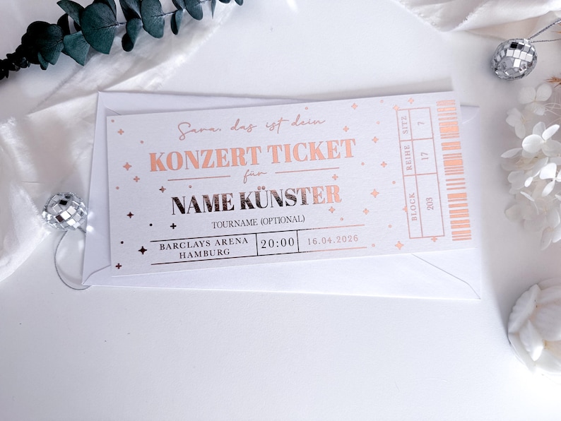Puede incluir: Una entrada de concierto con texto en oro rosa sobre fondo blanco. La entrada dice "KONZERT TICKET" y "NAME K&Uuml;NSTER" con el lugar y la fecha impresos debajo. La entrada est&aacute; dentro de un sobre blanco, con elementos decorativos.