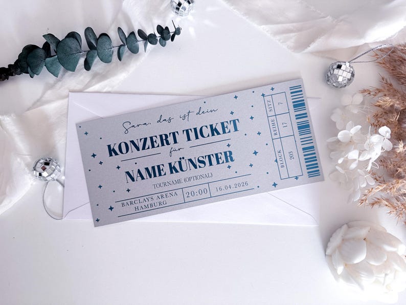 Puede incluir: Una entrada de concierto con texto azul y estrellas, dentro de un sobre blanco. La entrada dice "KONZERT TICKET" e incluye detalles del lugar, la fecha y la hora. Elementos decorativos incluyen bolas de discoteca y vegetaci&oacute;n.