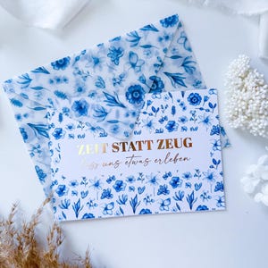 Könnte beinhalten: Ein Grußkarten- und Umschlag-Set mit blauem Blumenmuster. Die Karte trägt den Text "ZEIT STATT ZEUg lass uns etwas erleben" in goldfarbenen Buchstaben. Der Umschlag hat ein passendes Blumenmuster.