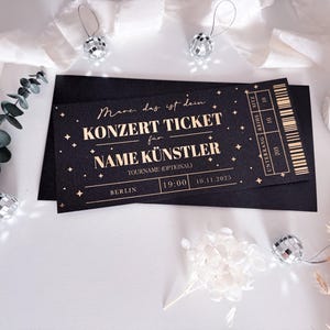 Puede incluir: Entrada de concierto negra con texto dorado, con "KONZERT TICKET" y "NAME K&Uuml;NSTLER". La entrada es para Berl&iacute;n, a las 19:00 el 10.11.2025. El dise&ntilde;o incorpora estrellas y un c&oacute;digo de barras, presentado en un sobre negro.
