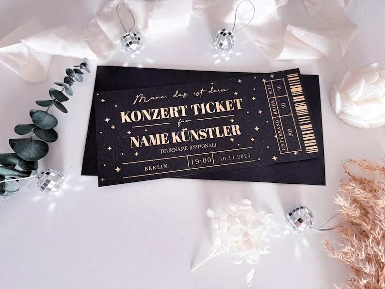 Puede incluir: Entrada de concierto negra con texto dorado, con "KONZERT TICKET" y "NAME K&Uuml;NSTLER". La entrada es para Berl&iacute;n, a las 19:00 el 10.11.2025. El dise&ntilde;o incorpora estrellas y un c&oacute;digo de barras, presentado en un sobre negro.