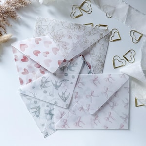 Puede incluir: Una colección de sobres decorativos con varios diseños, incluyendo corazones, flores, golondrinas y lazos. Clips dorados en forma de corazón y cinta blanca se encuentran alrededor de los sobres, creando una estética delicada y romántica.