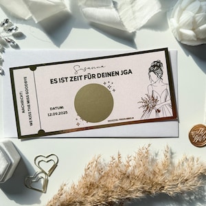 May include: A wedding-themed scratch card with the text "ES IST ZEIT FÜR DEINEN JGA" and "WE KISS THE MISS GOODBYE". The card features a bride illustration and the date 12.09.2025. Other items include a ring box, heart-shaped paper clips, and dried flowers.