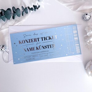 Puede incluir: Un boleto de concierto azul claro con texto plateado y estrellas, dentro de un sobre blanco. El boleto dice "KONZERT TICKET" y "NAME K&Uuml;NSTER". Elementos decorativos incluyen eucalipto, bolas de discoteca y flores secas.
