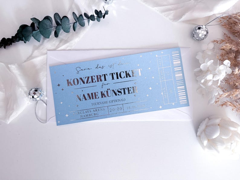 Puede incluir: Un boleto de concierto azul claro con texto plateado y estrellas, dentro de un sobre blanco. El boleto dice "KONZERT TICKET" y "NAME K&Uuml;NSTER". Elementos decorativos incluyen eucalipto, bolas de discoteca y flores secas.