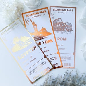 Könnte beinhalten: Drei Bordkarten mit Goldfolienakzenten. Reiseziele sind New York, Rom und Ägypten, mit dem Text "BOARDING PASS" und "ES GEHT NACH". Jede Karte hat ein Datum und einen Namen. Auf weißem Hintergrund.