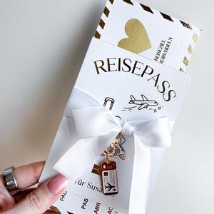 Könnte beinhalten: Eine weiße Karte im Reisepass-Design mit dem in Gold gedruckten Wort REISEPASS. Sie ist mit einer weißen Schleife versehen, an der ein kleiner rot-weißer Flugticket-Anhänger befestigt ist. Ein goldenes Herz ist am oberen Rand zu sehen.