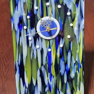May include: A rectangular glass clock featuring a vibrant, abstract pattern of vertical streaks in shades of blue, lime green, and white. The clock face has a central blue and white circular dial with gold-toned ornate hands and small white dots.