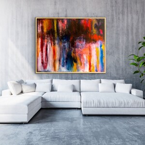 May include: Abstract painting with vertical brushstrokes in shades of red, orange, yellow, blue, and white. The painting is framed in gold and hangs on a gray wall above a white sectional sofa.