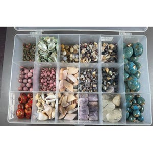 May include: A clear plastic storage container with fifteen compartments filled with various loose beads and natural elements. The collection includes green shell pieces, smooth round pink beads, white shell fragments, dark metallic chips, and teal speckled oval beads.