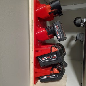 Op de afbeelding: Een houten wandbevestiging met rode kunststof houders voor het opbergen van vier accu's voor draadloze boormachines. De accu's zijn gelabeld met "M18 REDLITHIUM XC5.0".