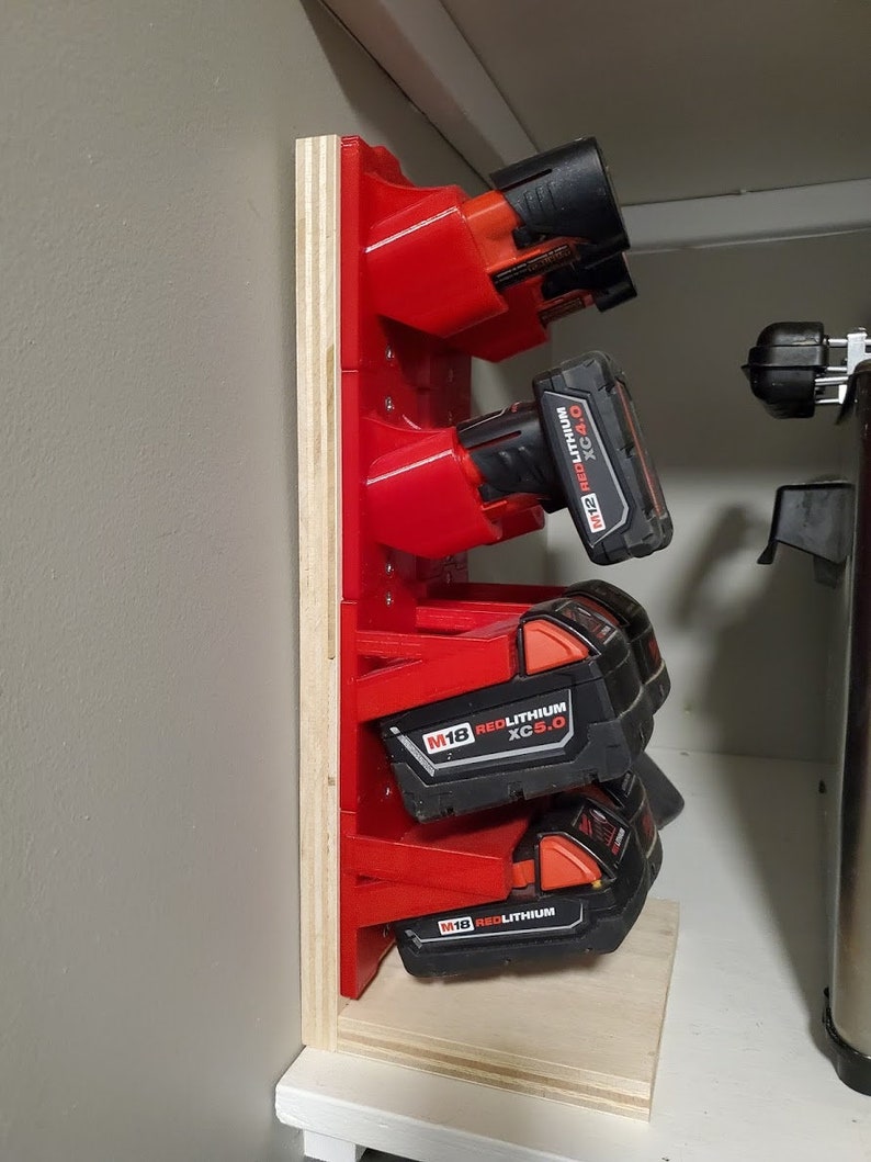 Op de afbeelding: Een houten wandbevestiging met rode kunststof houders voor het opbergen van vier accu's voor draadloze boormachines. De accu's zijn gelabeld met "M18 REDLITHIUM XC5.0".