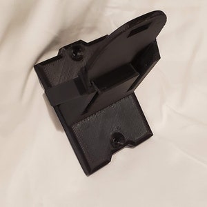 Op de afbeelding: Een zwarte 3D-geprinte wandbeugel met een gleuf voor een apparaat. De beugel heeft twee schroefgaten om aan een muur te bevestigen.