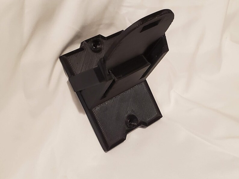 Op de afbeelding: Een zwarte 3D-geprinte wandbeugel met een gleuf voor een apparaat. De beugel heeft twee schroefgaten om aan een muur te bevestigen.