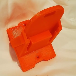 Op de afbeelding: Oranje 3D-geprint plastic wandbevestiging met een rechthoekige vorm en een klein gat aan de onderkant.