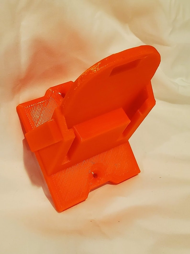 Op de afbeelding: Oranje 3D-geprint plastic wandbevestiging met een rechthoekige vorm en een klein gat aan de onderkant.