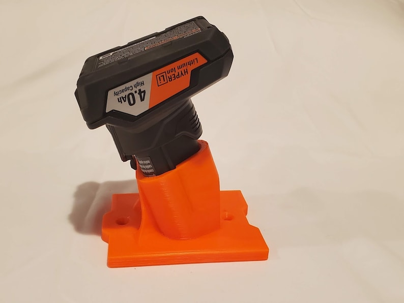 Op de afbeelding: Oranje 3D-geprinte wandbevestiging voor een zwarte Hyper Tough 4,0Ah lithium-ionbatterij. De beugel heeft twee gaten om aan een muur te bevestigen.