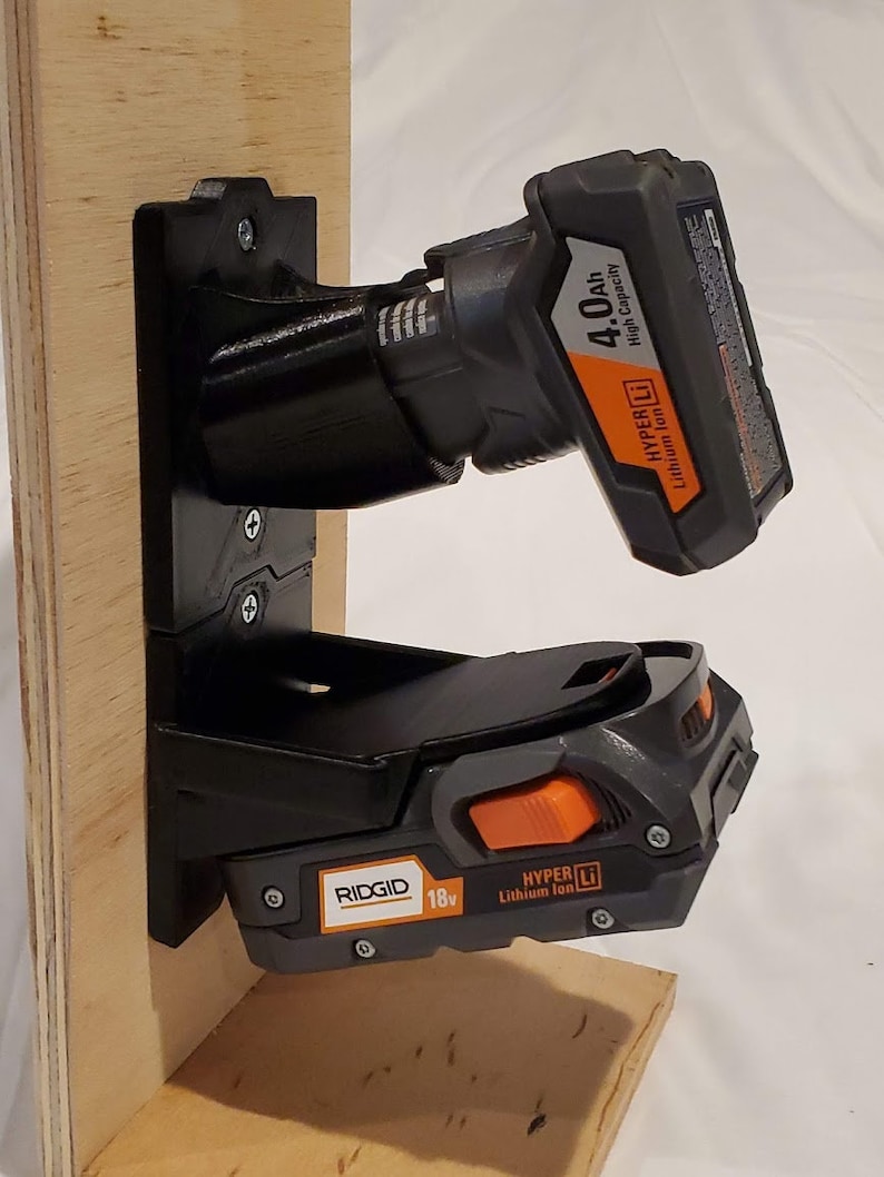 Op de afbeelding: Een zwarte wandbevestiging voor twee Ridgid 18v Hyper Lithium-ionbatterijen. De bevestiging is bevestigd aan een stuk hout. De batterijen bevinden zich beide in de bevestiging. De bovenste batterij is een 4,0Ah-batterij. De onderste batterij is een Hyper Lithium-ionbatterij.