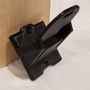 Op de afbeelding: Een zwarte 3D-geprinte wandbeugel met een rechthoekige basis en een gebogen arm die naar boven uitsteekt. De beugel heeft twee schroefgaten op de basis en &eacute;&eacute;n op de arm.