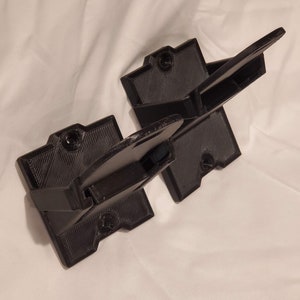 Op de afbeelding: Twee zwarte 3D-geprinte wandbeugels voor het ophangen van items. De beugels hebben een vierkante basis en een gebogen arm die naar buiten steekt.