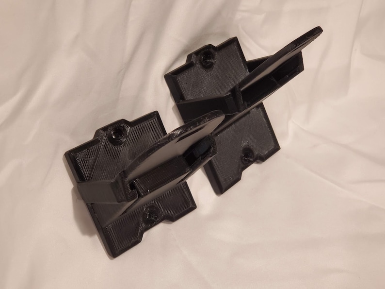 Op de afbeelding: Twee zwarte 3D-geprinte wandbeugels voor het ophangen van items. De beugels hebben een vierkante basis en een gebogen arm die naar buiten steekt.
