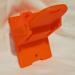 Op de afbeelding: Oranje plastic wandbeugel met een gleuf om een apparaat vast te houden. De beugel heeft twee gaten om aan een muur te bevestigen.