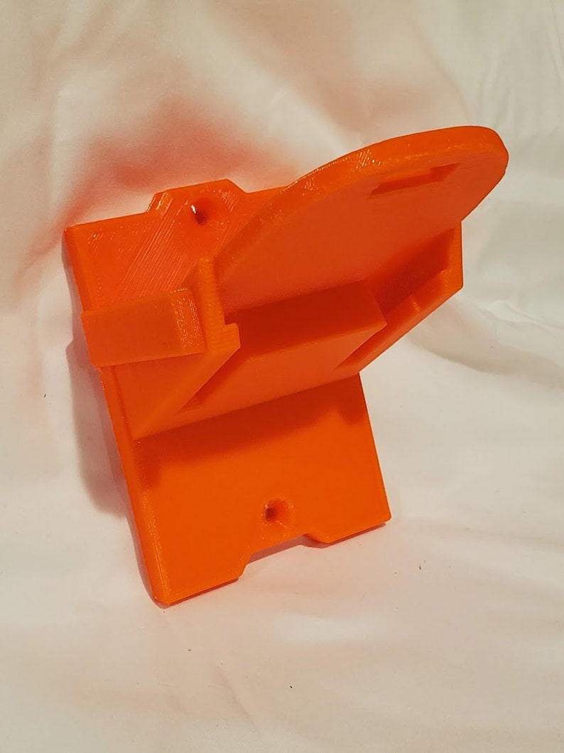 Op de afbeelding: Oranje plastic wandbeugel met een gleuf om een apparaat vast te houden. De beugel heeft twee gaten om aan een muur te bevestigen.