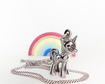 Könnte beinhalten: Eine silberne Halskette mit einem Einhorn-Anhänger und einer silbernen Kette. Der Anhänger ist eine detaillierte Darstellung eines Einhorns mit einem Horn und einem großen Auge. Die Halskette liegt auf einer weißen Oberfläche mit einem Regenbogen dahinter.