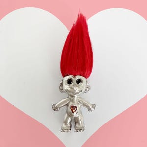 May include: A silver troll figurine with red hair and a red heart on its chest. The troll has black eyes and a small smile.
