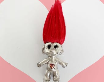May include: A silver troll figurine with red hair and a red heart on its chest. The troll has black eyes and a small smile.