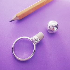 May include: A silver-toned ring featuring a cylindrical post topped with a small, light-coloured cylindrical insert, accompanied by a separate, detached, polished silver-toned spherical bead with a hole, positioned near a yellow wooden pencil.