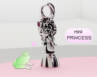Könnte beinhalten: Ein silberner Anhänger in Form einer Prinzessin mit einer Schlaufe oben. Die Prinzessin hat lockiges Haar, ein Kleid und einen roten Edelstein. Der Text "MINI PRINCESS" befindet sich in einem rosa Oval.