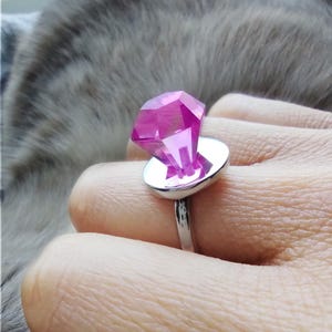 May include: A silver ring with a large, pink, faceted gemstone. The gemstone sits on a circular silver base, with a silver band. This statement ring is likely for fashion or costume purposes, and is a bold accessory.