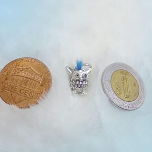 K&ouml;nnte beinhalten: Silberner Anh&auml;nger eines flauschigen blauen und wei&szlig;en Eulen mit einem blauen Irokesenschnitt. Der Anh&auml;nger befindet sich neben einem US-Penny und einem mexikanischen Peso.