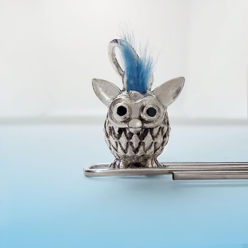 K&ouml;nnte beinhalten: Silbernes Lesezeichen mit einer kleinen, detaillierten, silbernen Figur eines pelzigen Wesens mit einem blauen Irokesenschnitt. Das Wesen hat gro&szlig;e, schwarze Augen und ein zahniges Grinsen. Das Lesezeichen ist an einem silbernen Metallclip befestigt.