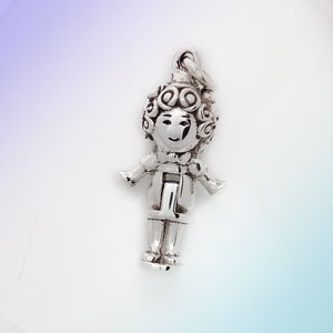 May include: Silver charm of a person with curly hair, wearing a jacket and holding a letter I.