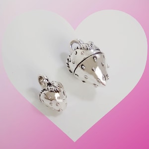 Puede incluir: Dos colgantes de plata con forma de fresa, con texturas detalladas y pequeños acentos transparentes. Los colgantes se muestran sobre un fondo blanco en forma de corazón con un degradado rosa.