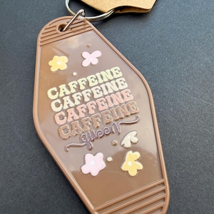 May include: A brown motel-style keychain with a metal ring and a cardboard tag. The keychain features the text CAFFEINE repeated four times in stacked rows, followed by the word queen in script, surrounded by small yellow and pink flower icons.