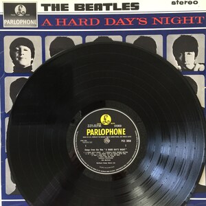 May include: A black vinyl record with a white label. The label has the text "The Beatles" and "A Hard Day's Night" in red and black lettering. The label also has the text "Parlophone" in black lettering and a small image of a pound sign.