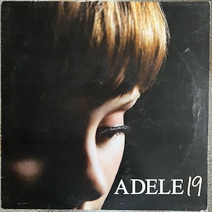 May include: Album cover featuring a close-up of a person's face with closed eyes and short, light brown hair. The album title "ADELE 19" is printed in white, lower right.