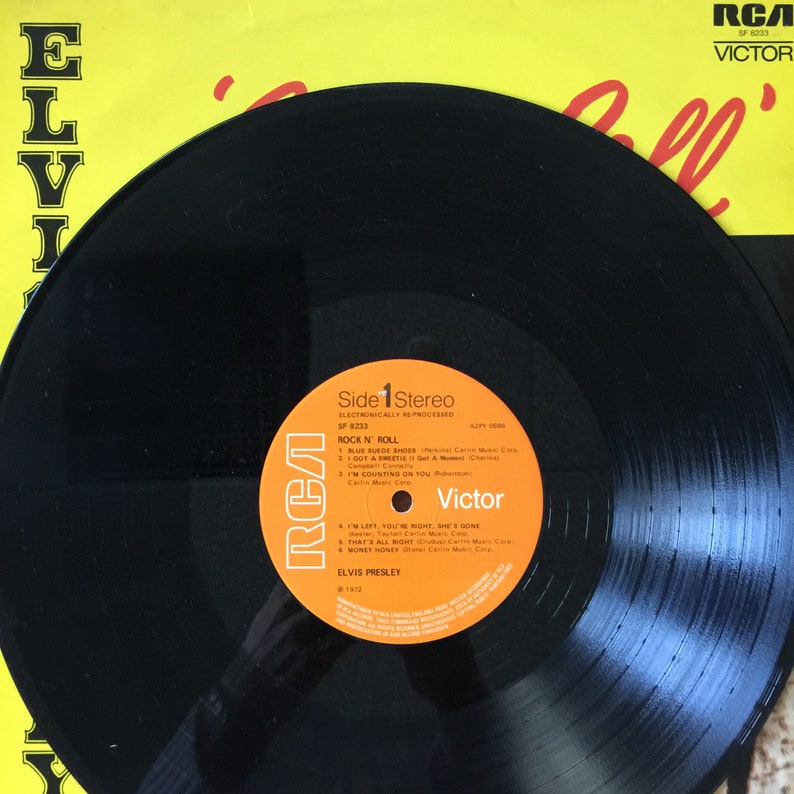 May include: A black vinyl record of Elvis Presley's "Rock N' Roll" album, with an orange and white label. The record is set against a yellow background with the RCA Victor logo. The album's tracklist is visible.