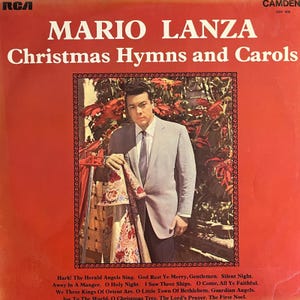 Pode incluir: Capa do álbum "Christmas Hymns and Carols" de Mario Lanza. A capa vermelha apresenta o nome do artista e o título do álbum em branco. Um homem de terno cinza posa. Os títulos das músicas são visíveis.