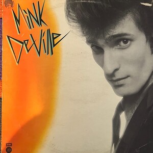 Puede incluir: Una portada de álbum de vinilo con una foto en blanco y negro de un hombre con cabello oscuro y traje. El título del álbum "Mink DeVille" está escrito en una fuente colorida y estilizada sobre un fondo naranja y amarillo brillante.