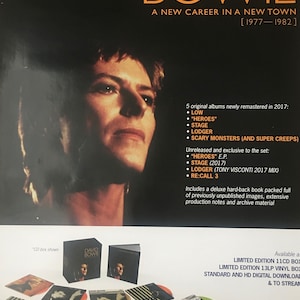 Op de afbeelding: Een zwart-witfoto van David Bowie, een muzikant, met de tekst "David Bowie A New Career in a New Town [1977-1982]" erboven. Onder de foto staat een lijst met albums en een beschrijving van een boxset met geremasterde albums en ongepubliceerd materiaal. De tekst "Out Now" staat onderaan de afbeelding.
