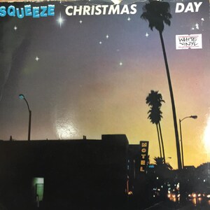 May include: A vintage vinyl record album cover featuring a nighttime scene with a motel sign, palm trees, and a starry sky. The album title is "Squeeze Christmas Day" with the words "White Vinyl" in a white box.